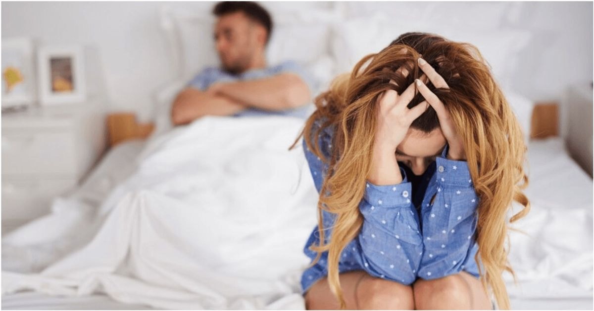 Nusivylusi liūdna mergina sėdi ant lovos susiėmusi už galvos, kai vyras tuo tarpu guli lovoje nepatenkintas